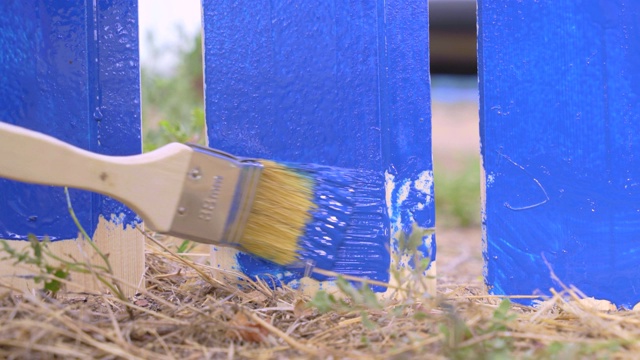 在村庄里，勤杂工用油漆刷和蓝色染料从木栅栏的木板底部油漆。地块内之维修及建设工作视频素材