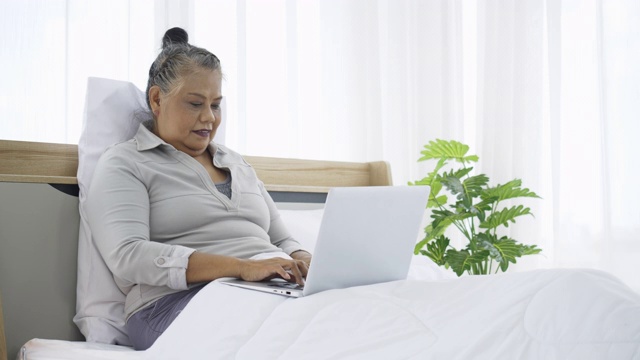 快乐的印度老女人坐在床上用笔记本电脑。网上购物的网站。病人在家通过远程保健使用笔记本电脑治疗疾病。虚拟医生预约视频素材