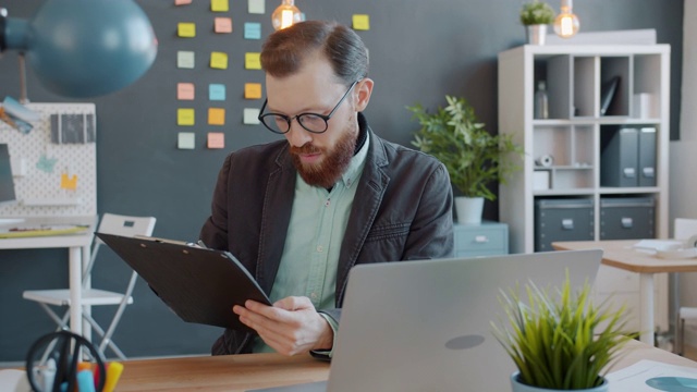 商人在创意办公室看着笔记本电脑屏幕做笔记的慢动作视频素材