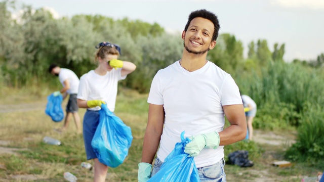 在镜头前，一个巨大的非洲裔美国人拿着一个蓝色的塑料袋，他和其他一些志愿者在海滩上捡垃圾视频下载