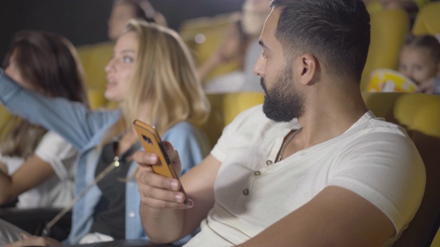 中东帅哥在电影院使用智能手机的侧视图，烦人的女性朋友在后面欢呼。无聊的家伙在电影院上网的肖像。视频素材