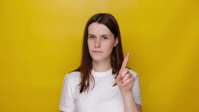 年轻的黑发女人的肖像警告用警告的手指手势，说不，给出建议，以避免危险，穿着白色t恤，不赞成的标志，孤立在黄色的工作室背景视频素材