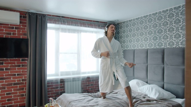 好玩的家伙穿着浴袍在床上跳舞通过耳机听音乐在酒店视频素材