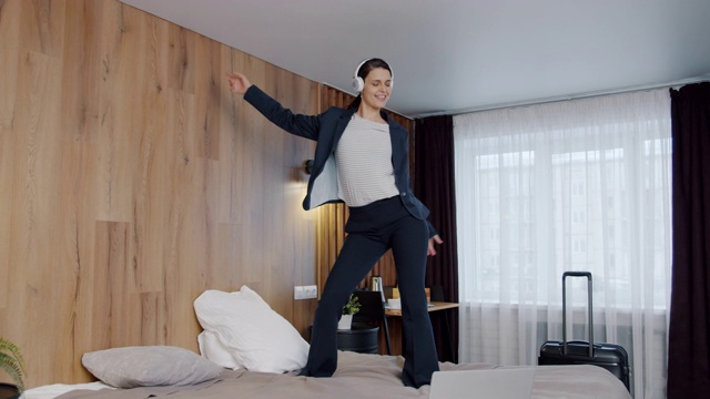 快乐的商务女士通过耳机听音乐在酒店的床上跳舞的慢动作视频素材