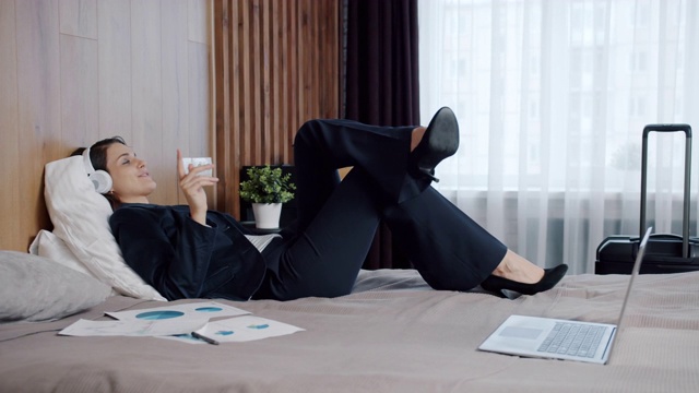 穿着西装的快乐女商人在酒店的床上通过无线耳机听音乐视频素材