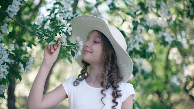 浪漫的女孩嗅着盛开的果园里的苹果树花。美丽的女孩戴着白色的帽子，穿着白色的衣服，在春天的花园里享受着樱花的芳香。视频素材