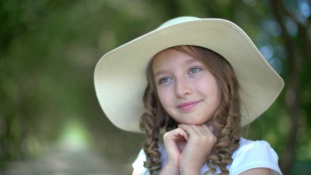 可爱的女孩肖像在白色的帽子牵手近脸在绿色的花园。可爱的年轻在夏天的帽子女孩微笑和看向一边在夏天的公园。视频下载