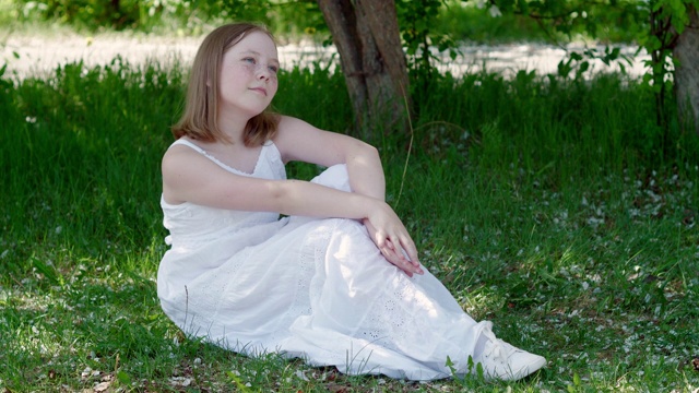 穿着白色连衣裙的轻松女孩坐在夏日花园的绿草地上。快乐的少女放松在绿色草坪在春天的果园。视频素材