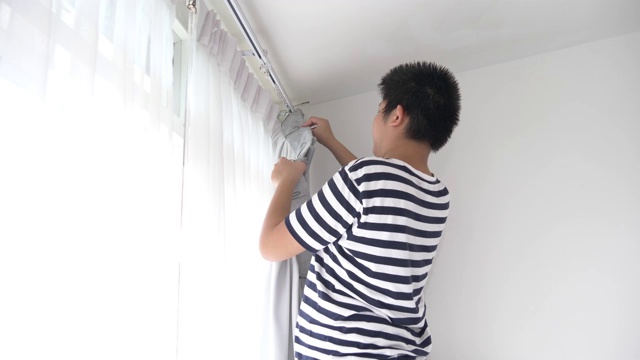 亚洲男孩在家里用窗户挂窗帘，搬家的概念。视频下载
