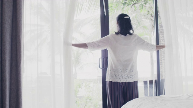 亚洲高级女性住在酒店房间里。打开房间的窗帘和门，看看外面的景色。假日旅游概念。4 k慢动作。视频素材