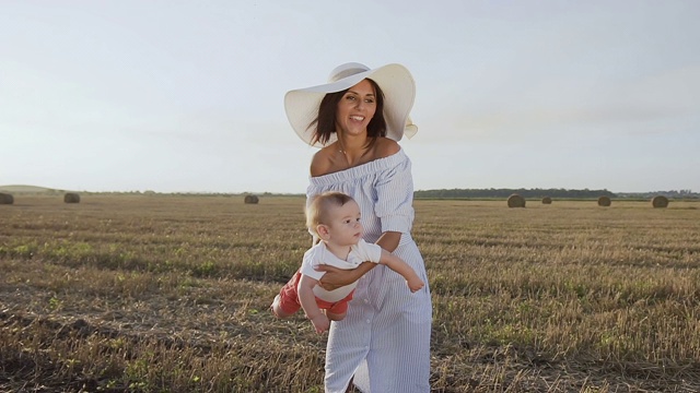 迷人的年轻女人穿着衣服和帽子是在日落的田野和她的孩子玩。夏天的一天，妈妈和儿子在地里玩着草包视频素材