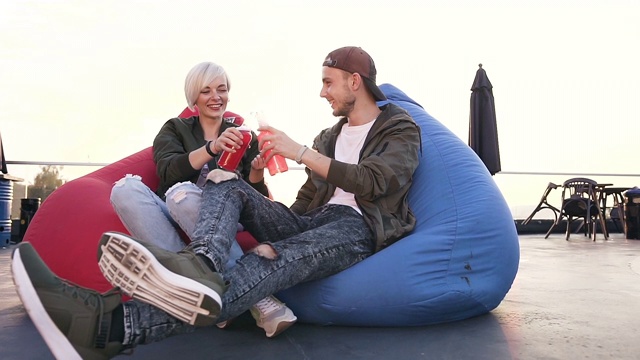 年轻迷人的时髦夫妇有乐趣坐在扶手椅袋与能量饮料的露台上的现代咖啡馆在日落。情绪激动、心情愉快的年轻人坐在五颜六色的扶手椅上，手里拿着饮料。在户外视频下载