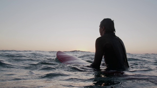 一个年轻人坐在他的冲浪板上等待日落的海浪视频素材