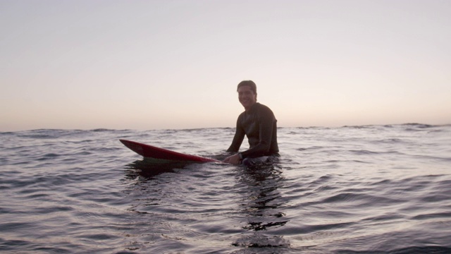 日落时分，一个年轻人坐在他的冲浪板上视频素材