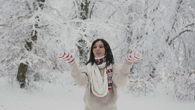 美丽的年轻女孩穿着羊毛衫和温暖的围巾，用她温暖的针织手套在雪地里捕捉雪花。一个女人从她的手套里吹雪花视频素材
