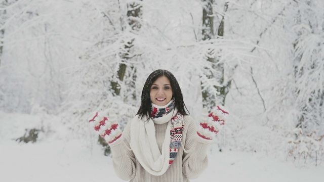 在白雪覆盖的森林里，一位年轻女子面带微笑，黑头发，穿着羊毛衫，围着温暖的围巾，戴着编织的连指手套，向人们挥手问好视频素材