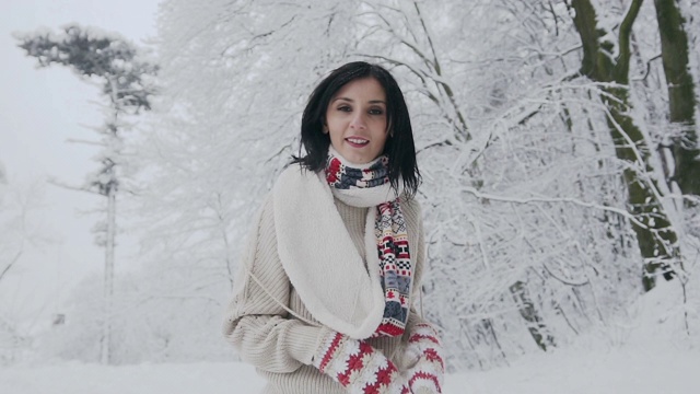 一个年轻的黑发女人的特写镜头在一个霜冻的冬天，谁走在白雪覆盖的森林在时尚的羊毛衫，温暖的围巾和针织手套视频素材