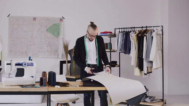 年轻的男性设计师与带子在他的脖子站在裁缝工作室和剪刀为一个新系列的模式。男胡子时装设计师和裁缝在工作室裁剪未来的衣服模式视频素材