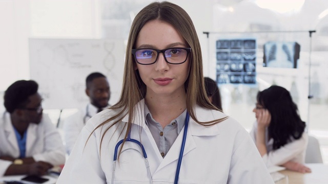 这是一幅自信的女医生的肖像，她戴着眼镜，身穿白大褂，脖子上戴着听诊器，对着医院同事身后的摄像机微笑。医生、保健、爱医的理念视频素材