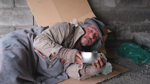 无家可归的老人躺在废弃建筑的地板上，拿着金属碗向其他人乞讨食物或钱的动作视频下载