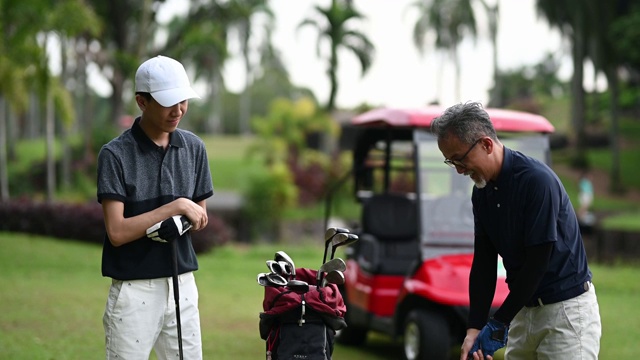 亚洲华人成年人从高尔夫球袋中挑选高尔夫球棒，并在高尔夫球场开球点指导教儿子视频素材