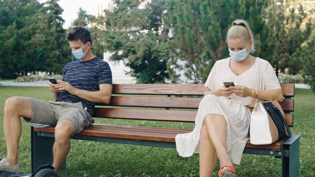 戴着防护口罩的情侣在公园的长椅上使用手机视频素材