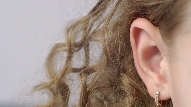 特写女性耳朵与耳环前视图。灰色背景上的卷发年轻女子的右耳。人类听觉器官。人体的部分。头部解剖。视频下载