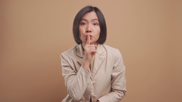 亚洲女商人把食指放在嘴唇上表示保守秘密，并站在米色背景下孤立。4 k的视频视频素材