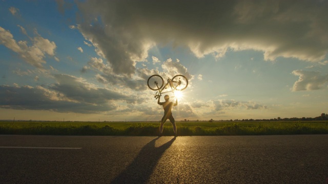 骑自行车的人站在路边，把自行车举过头顶。夕阳在背景中。4 k视频下载