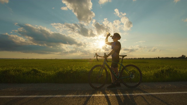 骑自行车的人站在路边，喝着瓶子里的水，背景是日落。照相机正把镜头对准他。4 k视频下载