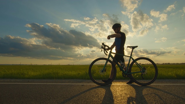 一个运动员骑着自行车沿着马路走。他正在看他的健身追踪器。骑自行车训练。绿色的田野和背景中的夕阳。4 k视频下载