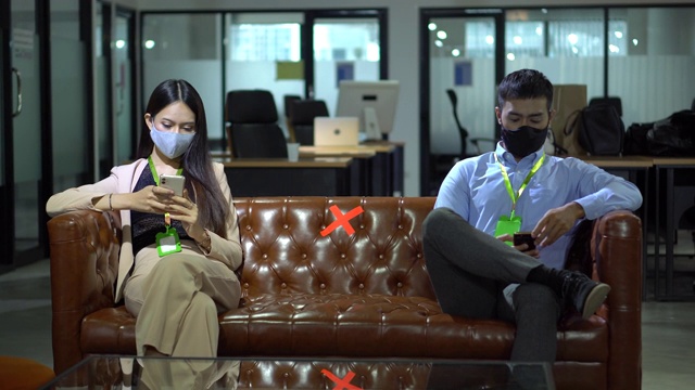 戴口罩的亚洲女员工和在办公室使用智能手机的商业人士与同事保持社交距离，以保护冠状病毒或covid-19疫情大流行。职场新常态政策视频下载