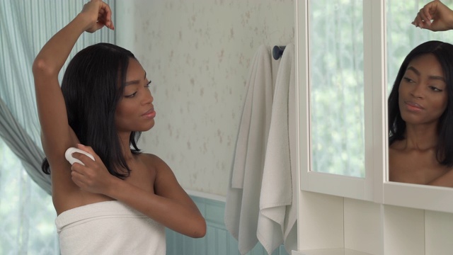 年轻的非洲妇女在腋下涂抹止汗剂，在浴室里照镜子。早晨例行公事。日常卫生的概念。视频素材