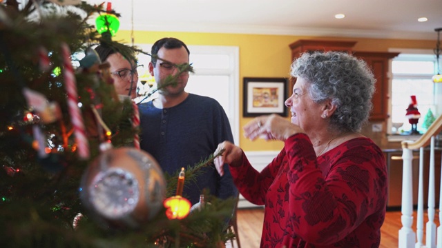 两代人的大家庭在平安夜在宽敞明亮的客厅里装饰圣诞树。当一对年轻夫妇在背景中交谈时，一位年长的女士正在调整装饰。视频素材