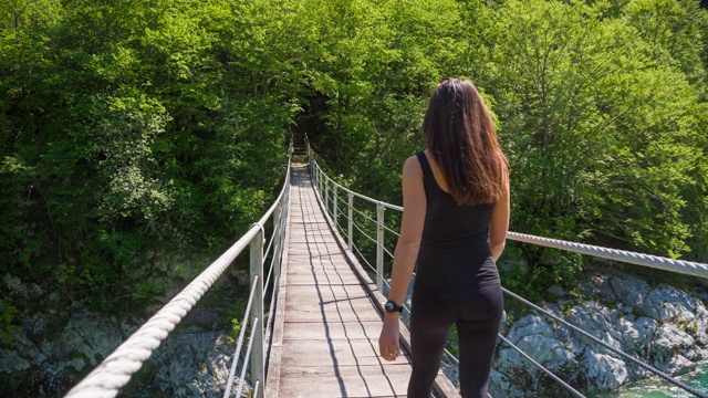 一名年轻女子正穿过山间溪流上的木吊桥视频素材