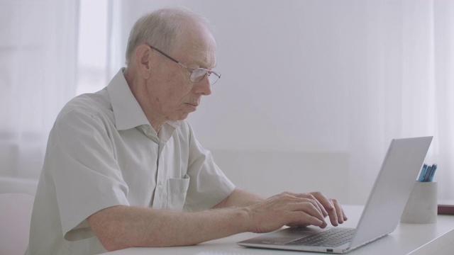 一位老人正在远程工作，通过电子邮件打字和发送信息，在笔记本电脑上上网，呆在家里视频下载