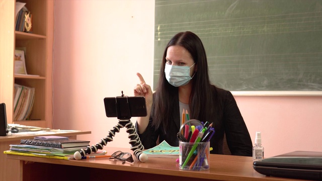 一位年轻的老师正在上课，用智能手机拍摄自己。女教师戴着防护口罩。隔离期间的视频教程视频素材