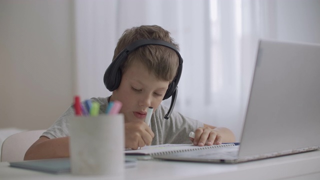 一个小男孩坐在家里的桌子旁，用耳机听音乐，用笔记本电脑画画视频下载