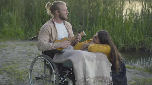 镜头接近微笑的残疾人在轮椅上弹奏尤克里里为美丽的年轻女子。正面的白人瘫痪男友与快乐的女朋友在日落户外约会肖像。视频素材