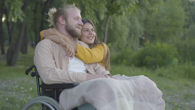 一幅沉思的年轻男子坐在轮椅上的肖像，就像一个快乐的美丽的女人跑向他和拥抱。快乐的残疾人男友和女友在夏夜约会。视频素材