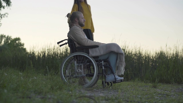 侧视图广角瘫痪的人在轮椅上休息与女朋友在户外黄昏。幸福的白人情侣在日落时分约会。残疾人的完整生活。视频素材