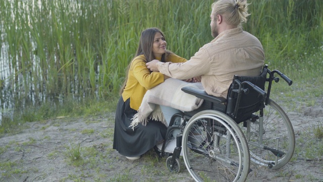 坐在轮椅上的白人截瘫男子，触摸着微笑的黑发女人。日落时分，快乐的女友和残疾的男友在河岸上约会。爱和浪漫。视频素材