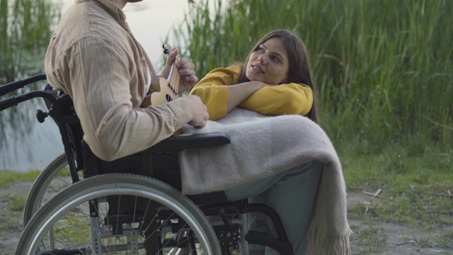 年轻可爱的女人听着轮椅上的残疾人弹奏四弦琴。美丽浪漫的白人女友与残疾男友在夏季日落户外约会的肖像。视频素材