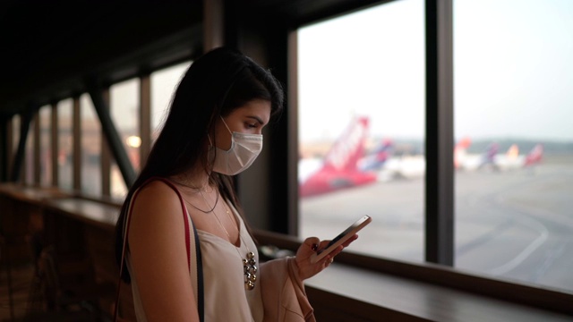 年轻的女旅行者透过窗户看，在机场使用电话-戴口罩视频素材