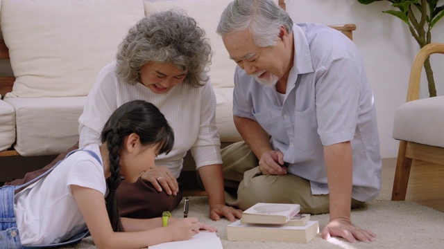 亚洲家族的祖父坐在客厅里笑着，用手摸着头表示爱。看孙女躺着写书，幸福老年退休视频下载