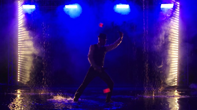 一个男人的剪影与发光的红色手榴弹在一个黑暗的烟雾演播室。一位时尚的艺人在雨中以蓝色的烟雾为背景进行火表演。慢动作视频素材