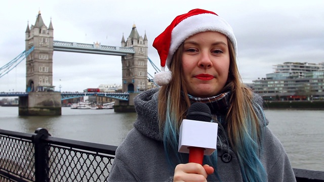 英国伦敦塔桥附近，年轻的女记者戴着圣诞帽，用麦克风讲话视频下载