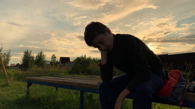 一个孤独悲伤的男孩坐在户外的长椅上，在夕阳下，不快乐的抑郁的孩子，有歧视和欺凌的问题视频素材