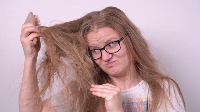 可爱的金发女孩戴着眼镜，试图用木刷梳理纤细纠结的长发，在白色的背景上打造时尚的发型，但没有成功。护理受损头发和头皮视频下载