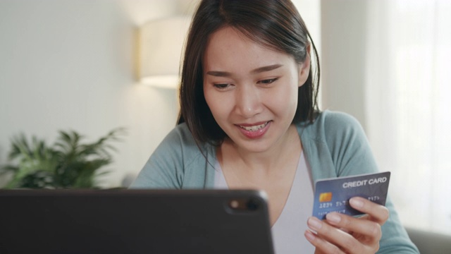 一位年轻漂亮的亚洲女性正在用信用卡进行在线支付，她坐在家里的起居室里，用平板电脑输入信用卡号码。网上购物，网上购物的概念。视频素材